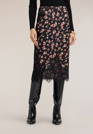 Kvinde iført en sort blomster-midi-nederdel med blondekant, sort ribbet langærmet top, guldarmbånd og sorte knæhøje støvler.