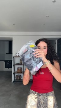 Juodos spalvos drabužiai skaidriame plastikiniame maišelyje su geltona kainų etiketė. Matomi ilgi garbanojami plaukai, raudona liemenė ir raštuoti apatiniai.
