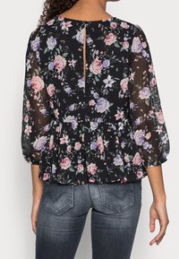 Schwarze Bluse mit Blumenmuster, langen, durchsichtigen Ärmeln, geraffter Taille und einem Schlüsselhole-Detail am Rücken. Mit pinken und blauen Blumemotiven auf einem dunklen Hintergrund.