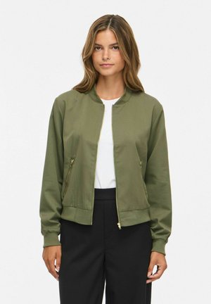 Femme portant une veste bomber kaki zippée sur une chemise blanche et un pantalon noir, debout devant un fond blanc uni.