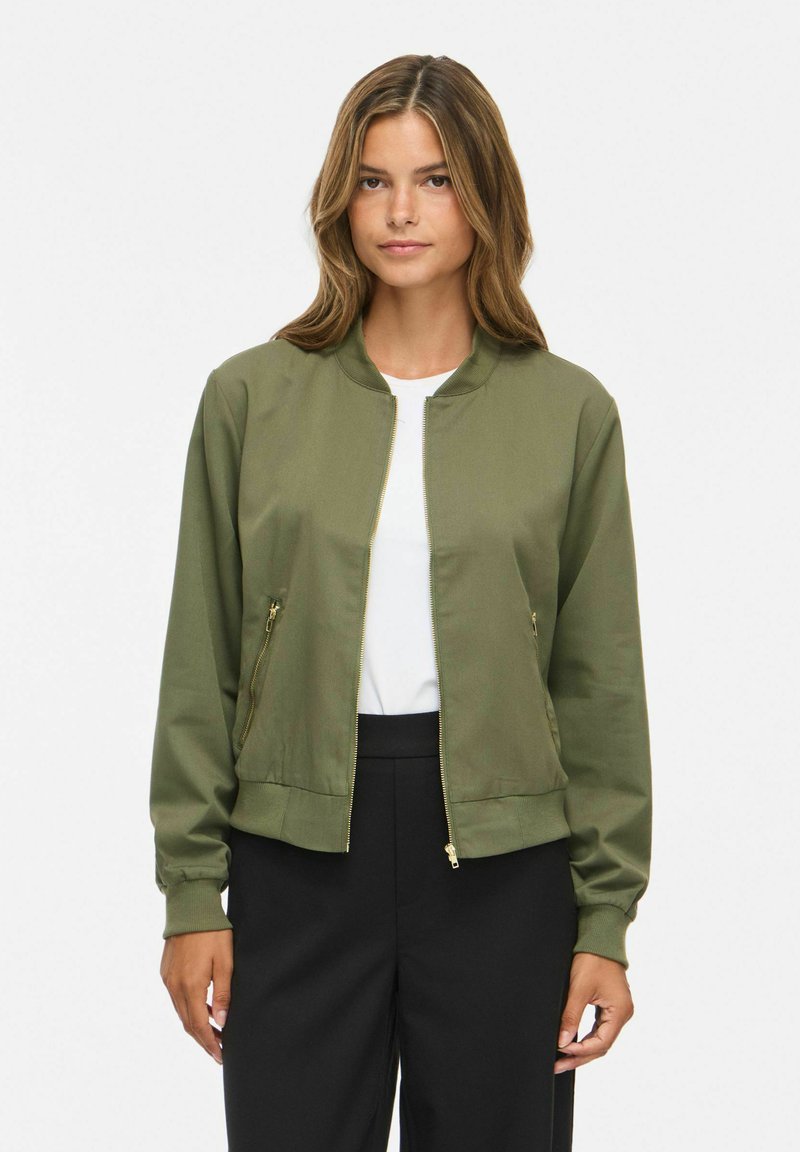 Femme portant une veste bomber kaki zippée sur une chemise blanche et un pantalon noir, debout devant un fond blanc uni.