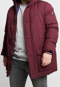 Giacca a vento color bordeaux con chiusura frontale a zip, grandi bottoni e tasche applicate. Polsini a costine con bordatura a contrasto. Interno foderato.