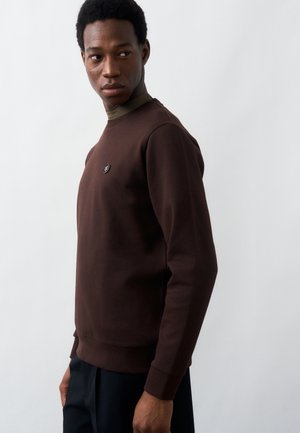 Άνδρας που φορά ένα σκούρο καφέ φούτερ με ένα μικρό στρογγυλό λογότυπο στο στήθος, στέκεται μπροστά σε απλό ανοιχτό φόντο, κοιτάζοντας πλάγια.