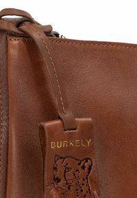 Angolo di una borsa in pelle marrone con finitura testurizzata, etichetta "BURKELY" in oro in rilievo e incisione a forma di leopardo su un cartellino appeso.