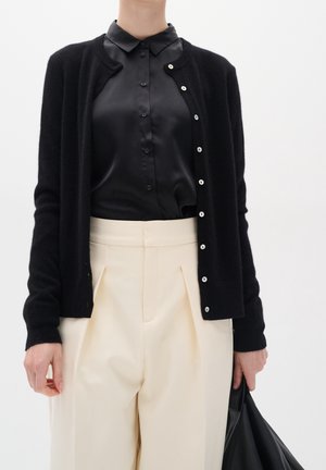 Kobieta w czarnym kardiganie na guziki, nosząca czarną satynową bluzkę, do tego kremowe, plisowane spodnie z wysokim stanem, trzymająca czarną torbę.