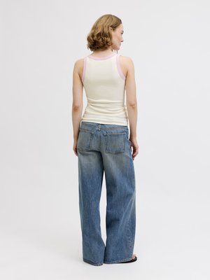 Femme aux cheveux courts et bouclés portant un débardeur blanc bordé de rose et un jean bleu ample, debout de dos sur un fond uni.
