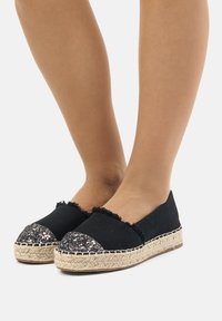 Espadrillas nere con tomaia in tessuto testurizzato, bordi scuciti e una punta nera luccicante. La suola avvolta in juta aggiunge un tocco naturale.