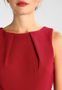 Robe sans manches rouge avec un décolleté rond, présentant deux plis à l'avant et un tissu lisse et texturé. Épaules et col sans coutures.