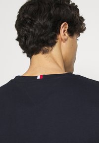 Námořnická modrá tričko s designem ve tvaru V, měkká látka a malý červený, bílý a modrý proužek na zadní části výstřihu.