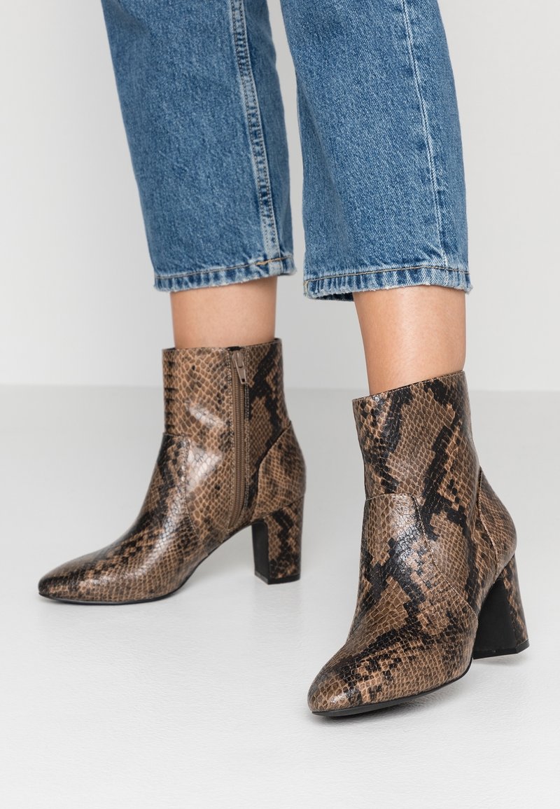 Brown snakeskin ankle boots with a pointed toe and block heel, featuring a side zipper and a glossy texture. Worn with cuffed blue jeans.