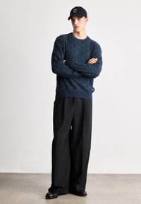 Marineblå genser med teksturert diamantmønster, kombinert med løse svarte bukser og svart caps med hvit tekst. Jevn stofffinish.