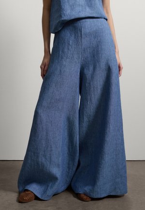 Donna che indossa pantaloni larghi in lino blu a vita alta abbinati a scarpe chiuse marroni intrecciate, in piedi davanti a uno sfondo semplice.