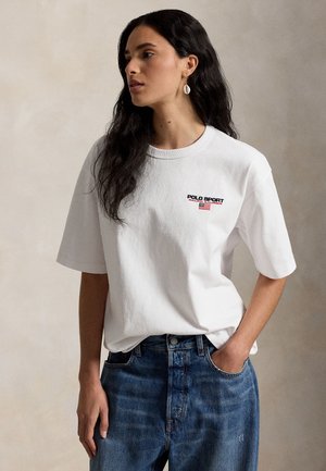 Γυναίκα με μακριά σκούρα μαλλιά, που φοράει λευκό T-shirt Polo Sport και μπλε τζιν, στέκεται με το ένα χέρι στην τσέπη, κοιτάζοντας αριστερά.