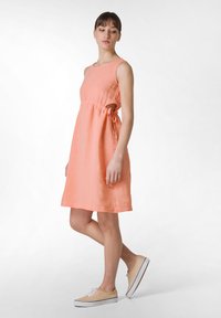 Persikofärgad linneklänning med en figursydd överdel, A-linjeformad silhuett, sidoutsprång och en knytning i midjan. Kombinerad med ljusa slip-on skor.