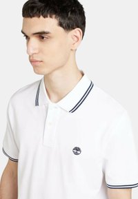 Polo bianco con colletto a costine con strisce blu navy, una patta con tre bottoni e un piccolo logo sul petto. Tessuto morbido.