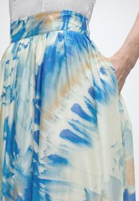 Φούστα με ψηλή μέση και μοτίβο δέσιμου σε μπλε και μπεζ αποχρώσεις ακουαρέλας, φορεμένη με λευκό τοπ που έχει μαζευτεί μέσα στη φούστα και χέρι ακουμπισμένο στην τσέπη της φούστας.