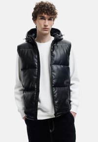 Schwarze Steppweste mit glänzendem Kunstleder-Finish, Reißverschluss und verstellbarem Kapuzen. Getragen über einem weißen Sweatshirt, mit schwarzen Hosen.