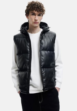 Musta puhvivesti kiiltävän tekonahkaisen pinnan kanssa, vetoketjukiinnitys ja säädettävä huppu. Vaatetuksessa valkoinen collegepaita ja mustat housut.