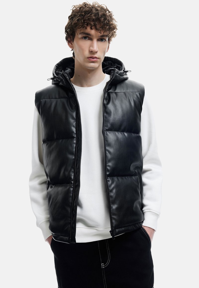 Schwarze Steppweste mit glänzendem Kunstleder-Finish, Reißverschluss und verstellbarem Kapuzen. Getragen über einem weißen Sweatshirt, mit schwarzen Hosen.