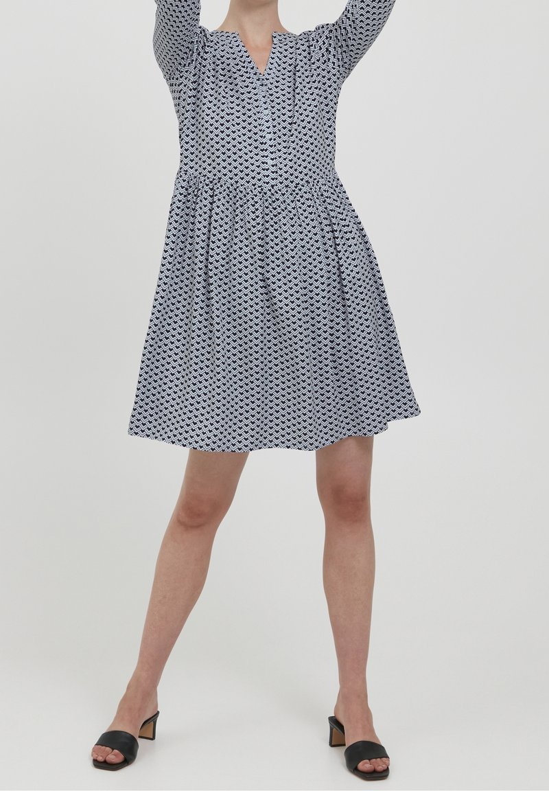 Femme portant une robe à motifs, longueur genou, manches longues, debout les bras levés, chaussée de sandales à talons noires.