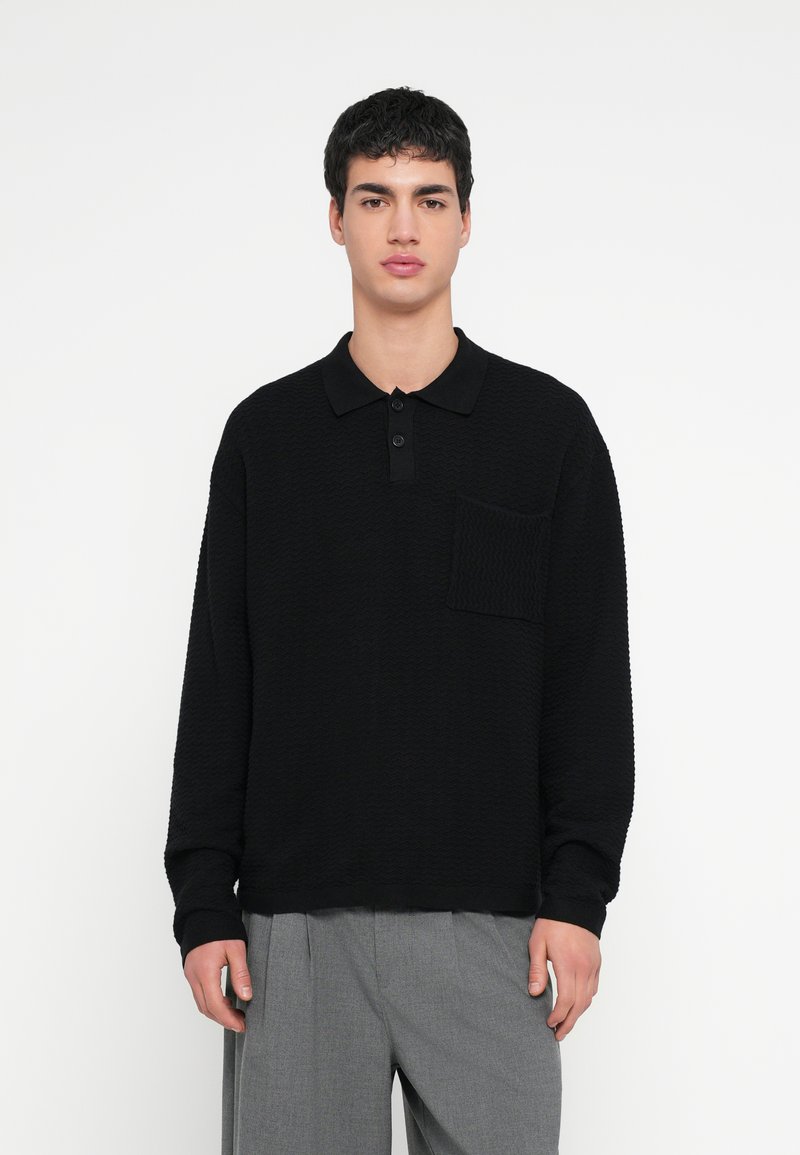 Μαύρο πόλο μπλουζάκι με μακριά μανίκια, υφή στο ύφασμα, τρία κουμπιά στη λαιμόκοψη και τσέπη στο στήθος. Φορεμένο με ανοιχτό γκρι παντελόνι.