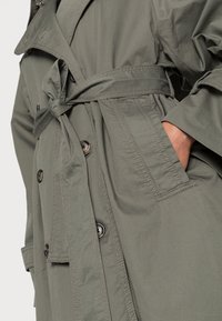 Khaki trencz z paskiem w talii, podwójnym zapięciem, dużymi guzikami oraz bocznymi kieszeniami, wykonany z trwałego, gładkiego materiału.