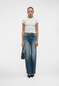 Ženska z opletenimi lasmi, oblečena v belo prilegajočo majico, široke modre kavbojke z visokim pasom in črne špicaste čevlje, drži črno torbico.