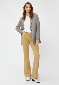 Grau karierter Blazer über einem weißen Oberteil, kombiniert mit beigen Schlaghosen und weißen Stiefeletten. Glatte Texturen und maßgeschneiderte Designs.