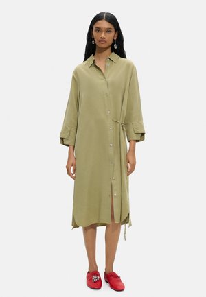Kobieta stojąca, ubrana w luźną oliwkowozieloną sukienkę zapinaną na guziki z rękawami 3/4 i czerwone buty sznurowane z ozdobnym srebrnym detalem.