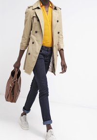 Beige trench coat s černými knoflíky, nošený přes jasně žlutou košili. V kombinaci s tmavými džíny a bílými šněrovacími botami. Hnědá kabelka držena v ruce.