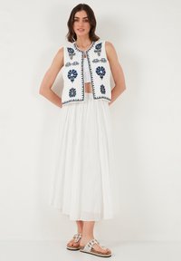 Valkoinen broderad liivi, jossa on sinisiä kukkakuoseja, yhdistettynä valkoiseen valuvaan hameeseen ja hopeisiin sandaaleihin, joissa on koristeellisia yksityiskohtia.