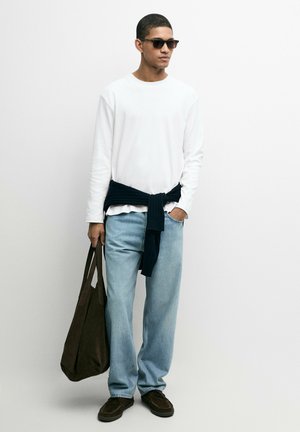 Uomo con occhiali da sole, camicia bianca a maniche lunghe, jeans azzurri, maglione scuro legato in vita, tiene una grande borsa marrone, in piedi contro un muro bianco.
