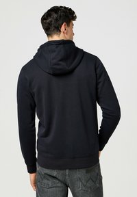 Black hoodie with a drawstring hood, fitted cuffs, and ribbed waistband. Soft fabric texture. Back view, worn with dark jeans featuring a logo patch.