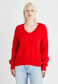Un pull tricoté rouge et duveteux avec un col en V et de longues manches légèrement bouffantes. Ourlet côtelé avec une coupe décontractée. Associé à un jean en denim bleu clair.