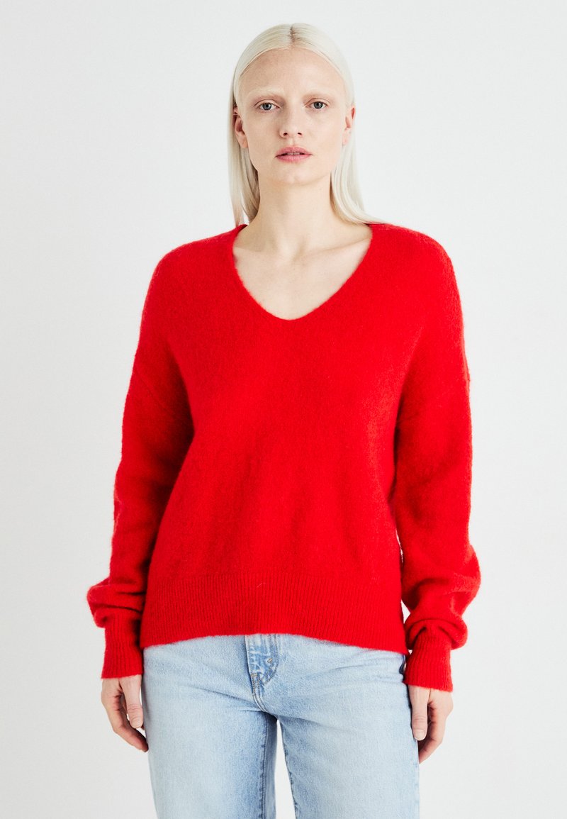 Un pull tricoté rouge et duveteux avec un col en V et de longues manches légèrement bouffantes. Ourlet côtelé avec une coupe décontractée. Associé à un jean en denim bleu clair.
