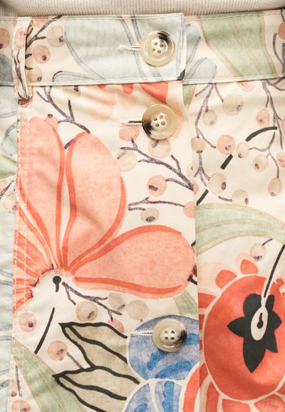 Κοντινή λήψη υφάσματος με μεγάλα λουλούδια σε ροδακινί, κόκκινο και μπλε χρώμα, μπεζ κουμπιά που κλείνουν και ορατές ραφές και ραμμένα σημεία.