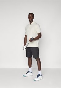 Polo shirt beige clair avec un col, shorts noirs, baskets blanches avec des accents bleu marine, tenant un club de golf, portant un gant de golf.