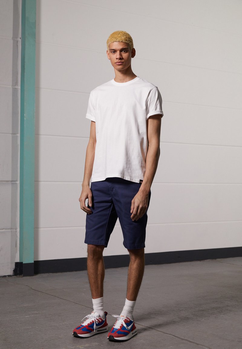 T-shirt de algodão branca, calções azul-escuro, sneakers vermelhos e azuis com detalhes brancos. Mangas curtas, gola redonda. Textura suave, design casual.
