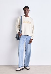Krém színű pulcsi, arany betűkkel a "BARBOUR INTERNATIONAL" felirattal, világoskék bő szabású farmerral és fekete talpú krém színű cipővel párosítva.