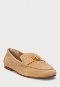 Beige suède loafers met een platte hak, ronde neus en een gouden accentlogo aan de voorkant. Glad textuur en gestikte details aan de zijkanten.