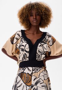 Korte mouwen blouse in beige met een zwarte bloemenprint, met een diepe V-hals en brede mouwen, accentueert met een zwarte tailleband.