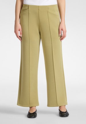 Donna che indossa pantaloni larghi color oliva chiaro con cuciture frontali e tasche laterali, abbinati a ballerine nere e una maglietta bianca.