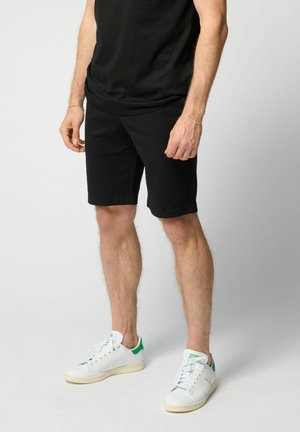 Muž oblečený v čiernych šortkách, čiernom tričku a bielych teniskách so zelenými detailmi, stojaci na jednolatom svetlom pozadí.