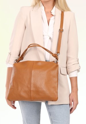 Woman wearing beige blazer and light jeans carrying a large brown leather handbag with shoulder and hand straps.