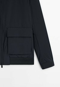 Manche de veste noire et partie inférieure avant avec grande poche à rabat et ourlet côtelé sur fond blanc.