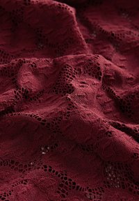 Tessuto in pizzo color bordeaux con motivi floreali intricati e superficie texture, che mostra trame aperte e caratteristiche di morbido drappeggio.