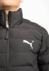 Czarna pikowana kurtka z wysokim kołnierzem, z białym logo Pumy na lewej piersi. Widoczne zapięcie na zamek i przeszycia.