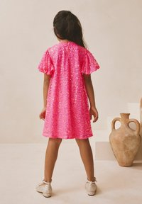 Rózsaszín flitteres ruha rövid, fodros ujjakkal és kerek nyakkivágással. A ruha térd fölé ér, csillogó, lapos cipővel párosítva.