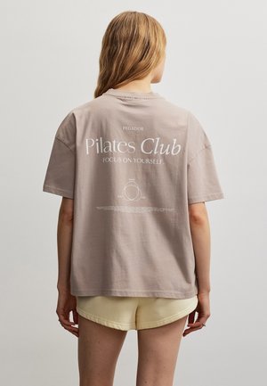 Kvinde med langt blondt hår iført en beige "Pilates Club" t-shirt og lysegule shorts, står med front mod en ensfarvet lys baggrund.