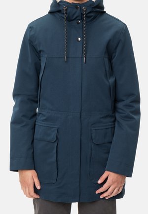 Hombre con chaqueta azul con capucha, botones delanteros, cordones y cuatro bolsillos, de pie frente a un fondo liso.